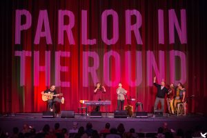 Four musicians on a stage with a red curtain behind them and the the words parlor in the round.