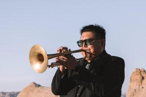 Man playing a trumpet against a backdrop of blue sky and rocks.